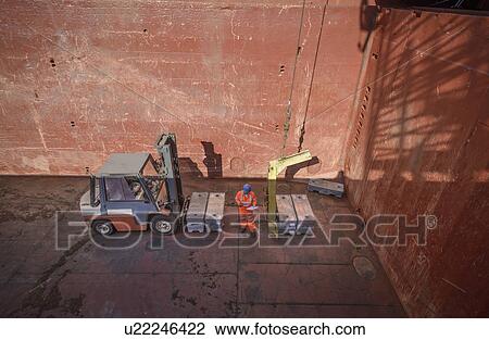 Stock Image - High angle view of worker checking metal alloy ingots in ship's hold. Fotosearch