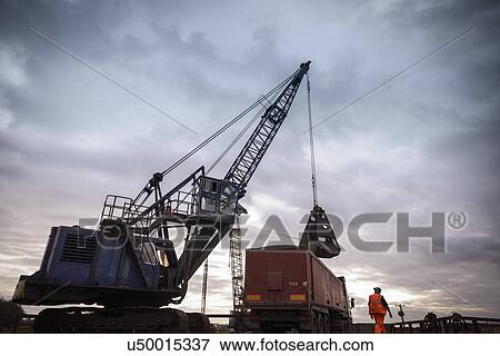 Stock Photo - Low angle view of crane loading truck under dramatic sky. Fotosearch