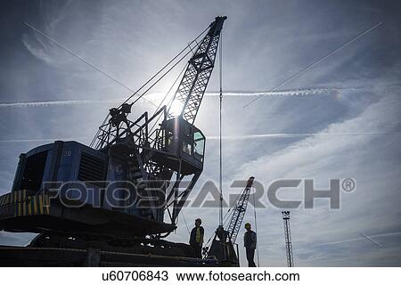 Stock Image - Low angle view of workers and crane in silhouette in port. Fotosearch