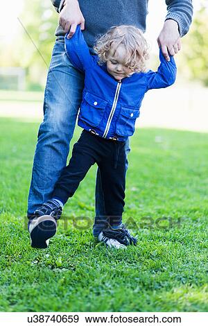 Stock Photo - Male toddler walking on fathers feet in park. Fotosearch