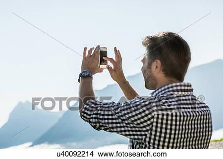 Picture - Man photographing view on his mobile phone, Tyrol, Austria. Fotosearch