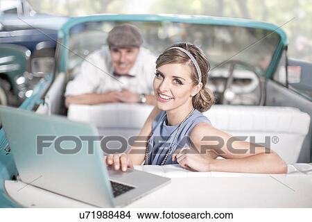 Picture - Man watching woman use laptop in back seat of convertible. Fotosearch