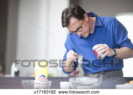 Stock Image - Mature man adding spoonful of baking powder to mixing bowl. Fotosearch