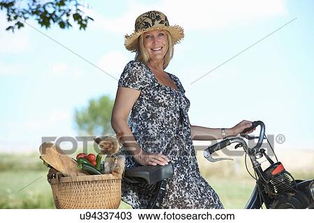 Stock Image - Mature woman on electric bike with dog and vegetables in basket. Fotosearch