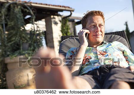 Stock Image - Mid adult man lying on sun lounger on phonecall. Fotosearch