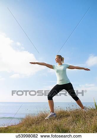 Stock Image - Mid adult woman stretching on cliff top, Thurlestone, Devon, UK. Fotosearch