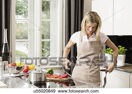 Stock Photo - Mid adult woman tying apron in kitchen. Fotosearch
