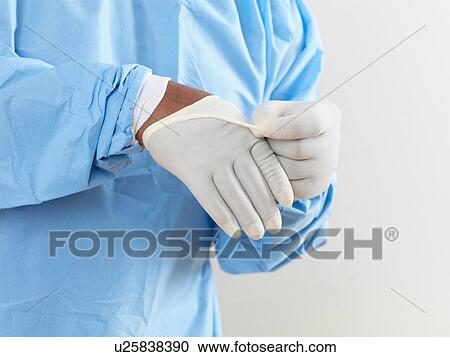 Stock Image - Mid section of surgeon wearing surgical scrubs putting on latex gloves. Fotosearch