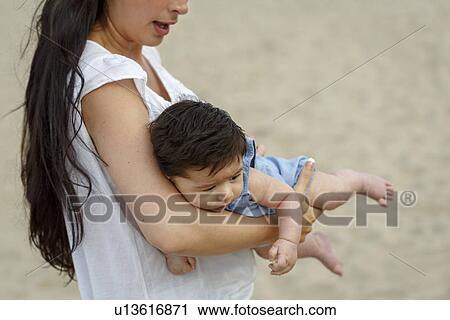 Stock Image - Mother on beach carrying baby in arms. Fotosearch