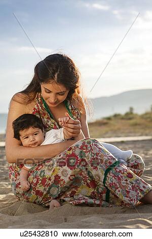 Stock Image - Mother sitting on sand, holding baby. Fotosearch