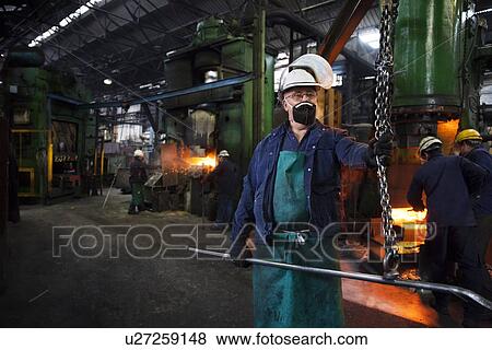 Stock Photo - Operative prepares to hook steel billet out of furnace. Team in background using counterblow hammer. Fotosearch