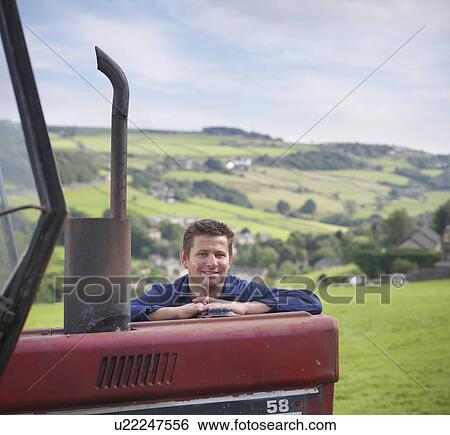 Stock Photograph - Portrait of farmer leaning on tractor in field, smiling. Fotosearch