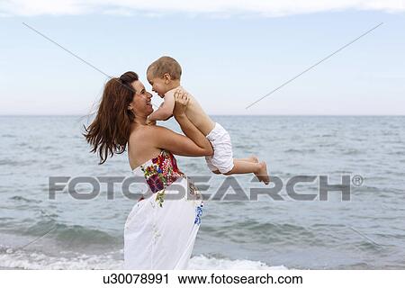 Stock Image - Pregnant woman lifting toddler on beach. Fotosearch