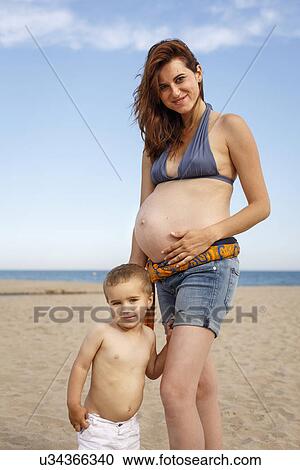 Stock Image - Pregnant woman on beach, hand on stomach, with toddler. Fotosearch