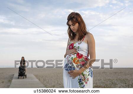 Picture - Pregnant woman standing on boardwalk, woman pushing pushchair in background. Fotosearch