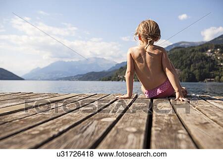 Stock Photo - Rear view of girl sitting on pier looking at view of lake and mountains. Fotosearch
