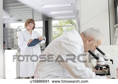 Stock Image - Scientists working in laboratory, man looking through microscope and woman taking notes. Fotosearch