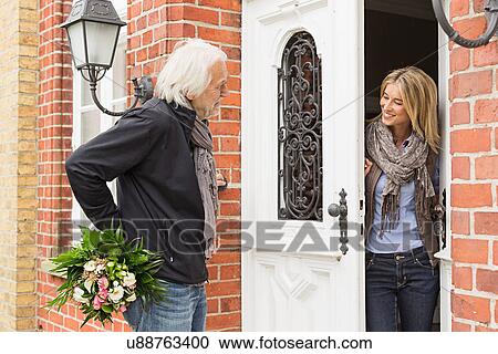 Stock Image - Senior man with bouquet, mid adult woman opening front door. Fotosearch