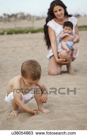 Stock Image - Toddler digging in sand with hand, mother and baby in background. Fotosearch