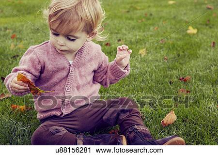 Stock Image - Toddler sitting on grass looking at autumn leaf. Fotosearch