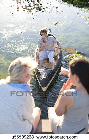 Two female friends sitting on pier pointing at young man rowing View Large Photo Image Stock Photo - Two female friends sitting on pier pointing at young man rowing. Fotosearch