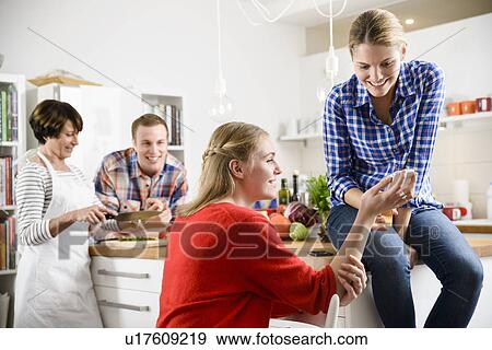 Stock Photo - Two women with cell phone in kitchen, mother and brother in background. Fotosearch