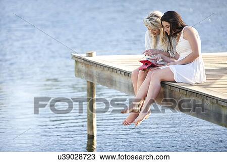 Stock Image - Two young women sitting on pier looking at digital tablet. Fotosearch