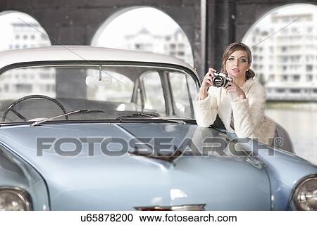 Stock Image - Woman leaning on convertible holding camera. Fotosearch