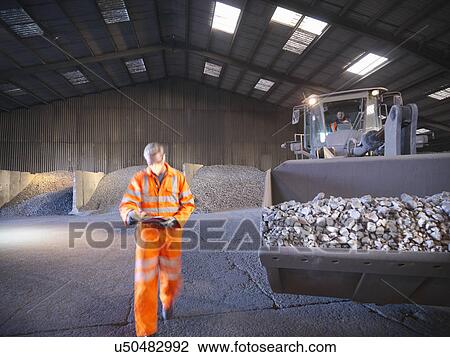 Stock Image - Worker checking clipboard in mineral store passing digger full of metal alloy. Fotosearch