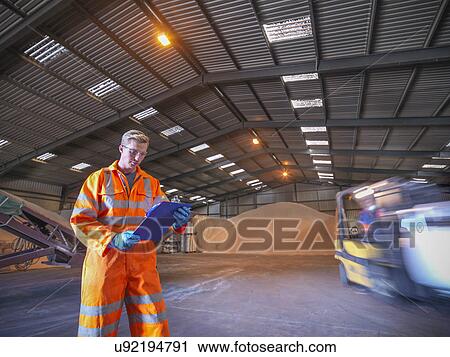 Stock Image - Worker checking clipboard in warehouse with zircon sand as forklift truck passes. Fotosearch