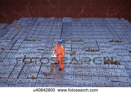 Stock Image - Worker in reflective workwear standing on metal alloy cargo in ship's hold. Fotosearch