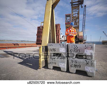 Stock Image - Worker inspecting metal alloy unloaded from ship. Fotosearch