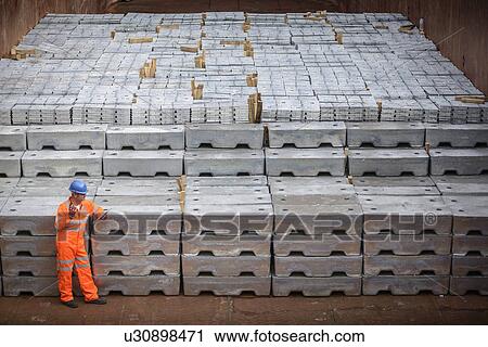 Stock Image - Worker standing next to metal ingots in ship's hold. Fotosearch