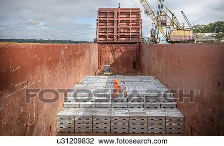 Stock Image - Worker standing on metal ingots in ship's hold. Fotosearch