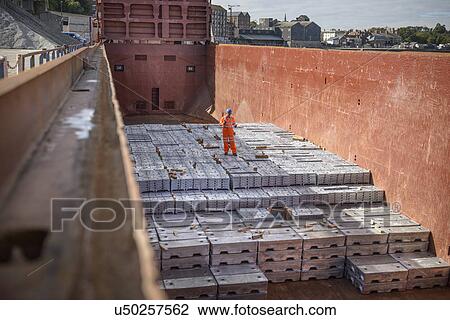 Stock Image - Worker standing on metal ingots in ship's hold. Fotosearch