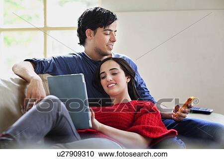 Stock Image - Young couple with digital tablet and mobile on living room sofa. Fotosearch