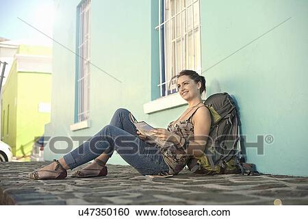 Stock Image - Young woman sitting outside blue painted house, reading map. Fotosearch