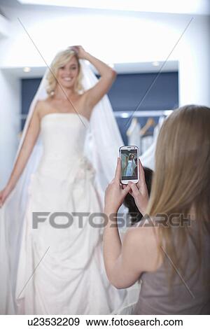 Young woman taking photograph of friend trying on wedding dress View Large Photo Image Stock Photo - Young woman taking photograph of friend trying on wedding dress. Fotosearch