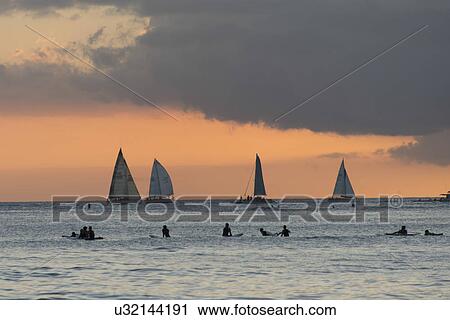 Stock Image - Surfers on the beach with sailboats in the background at sunset, Waikiki, Honolulu, Oahu, Hawaii, USA. Fotosearch