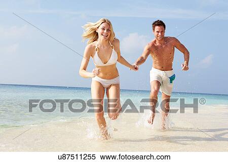 Stock Photography - Couple Running Through Waves On Beach Holiday. Fotosearch