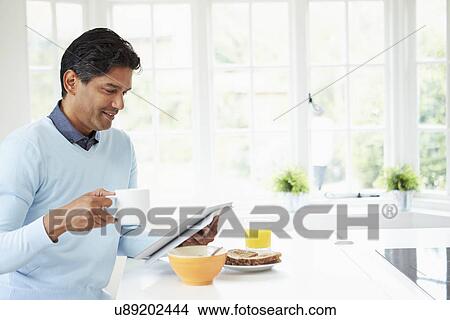 Picture - Indian Man Using Digital Tablet Whilst Eating Breakfast. Fotosearch