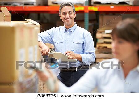 Stock Image - Manager In Warehouse With Worker Scanning Box In Foreground. Fotosearch