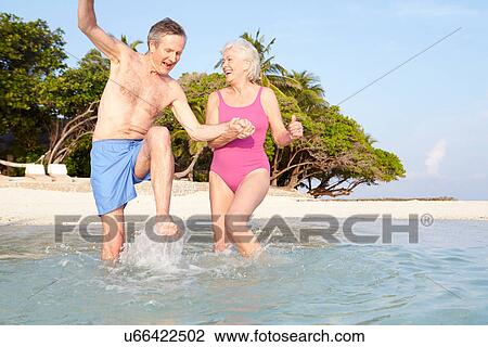 Stock Image - Senior Couple Splashing In Sea On Tropical Beach Holiday. Fotosearch