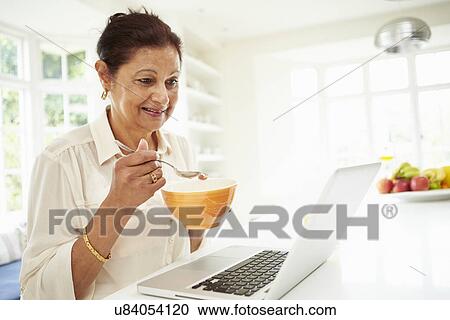 Stock Image - Senior Indian Woman Using Laptop Whilst Eating Breakfast. Fotosearch