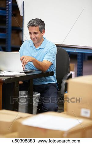 Stock Image - Worker In Distribution Warehouse Using Laptop. Fotosearch