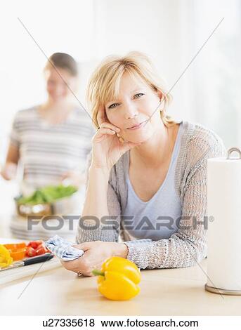 Stock Photo - Mother leaning on kitchen counter, out-of-focus daughter in background. Fotosearch