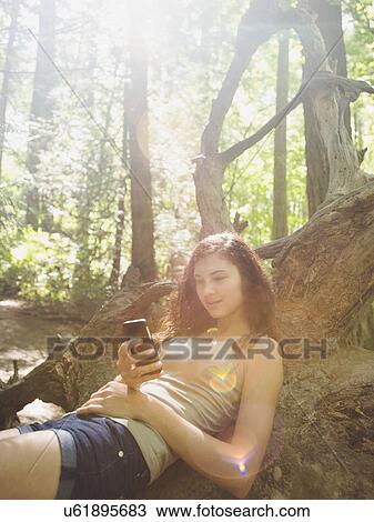 Stock Image - Young woman sitting on log and text messaging. Fotosearch