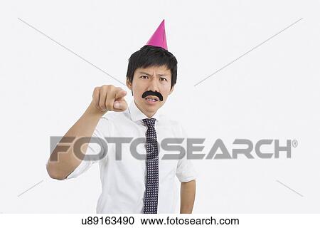 Stock Image - Angry businessman wearing party hat pointing in office. Fotosearch