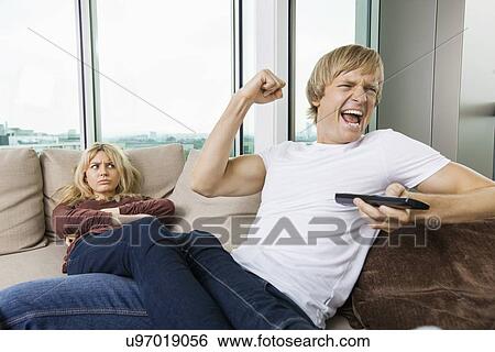 Angry woman staring at cheerful man as he watches TV in living room at home View Large Photo Image Stock Photograph - Angry woman staring at cheerful man as he watches TV in living room at home. Fotosearch