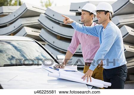 Stock Image - Architects with blueprints on car discussing at site. Fotosearch